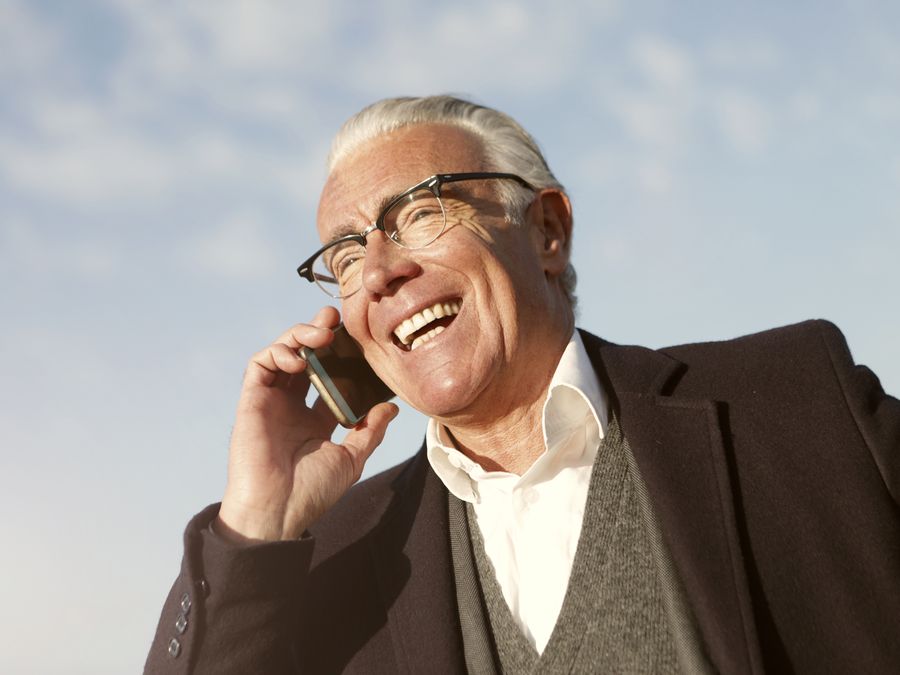 Happy senior man wearing glasses laughing while talking on smartphone outside.