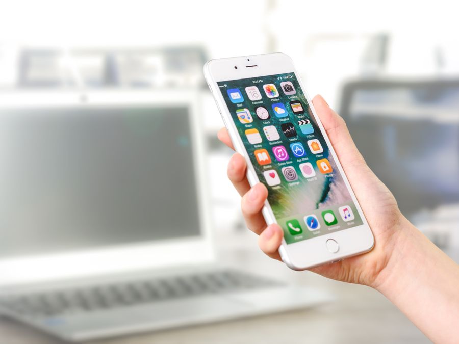 Close-up view of a smartphone displaying apps, held by a hand, with a blurred laptop in the background.