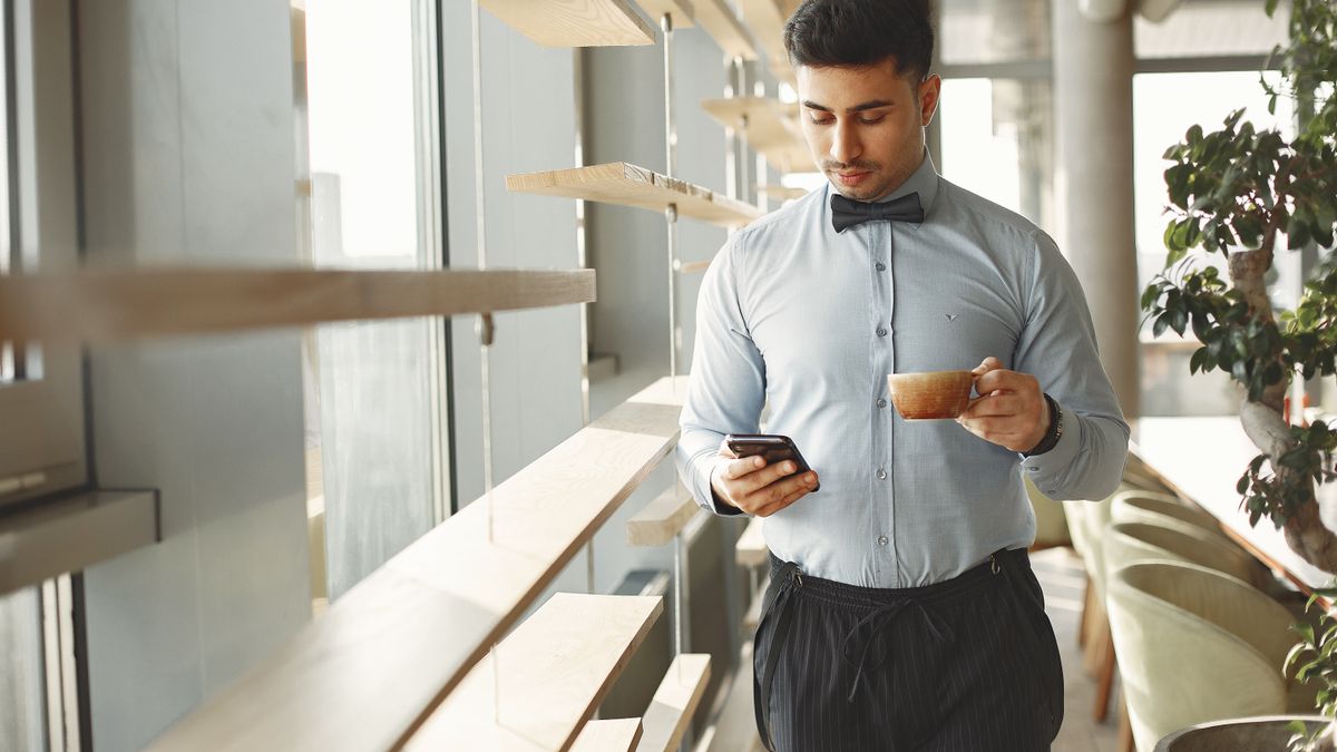 Man in business attire holding coffee cup, using smartphone in modern cafe.