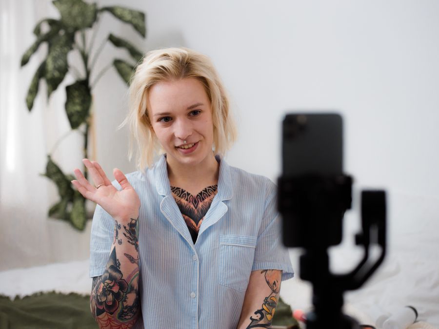 Tattooed woman with blonde hair smiling and recording video using a smartphone indoors.