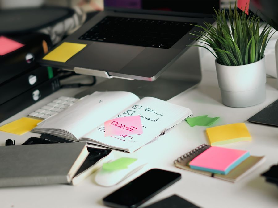 A cluttered office desk featuring a laptop, notebooks, and checklists amid colorful sticky notes.