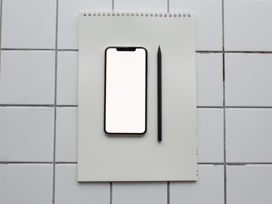Flat lay of a smartphone with blank screen on a notebook next to a pencil, on a tiled surface.