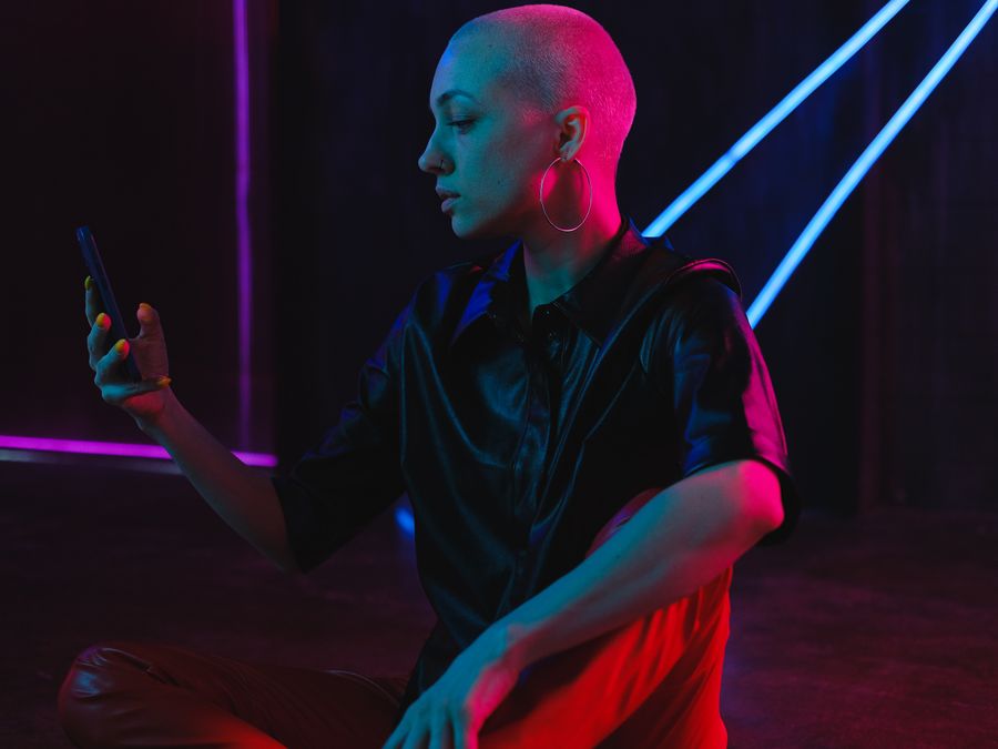 Pensive bald female in black leather shirt browsing modern mobile phone and sitting on floor in neon lights in dark studio