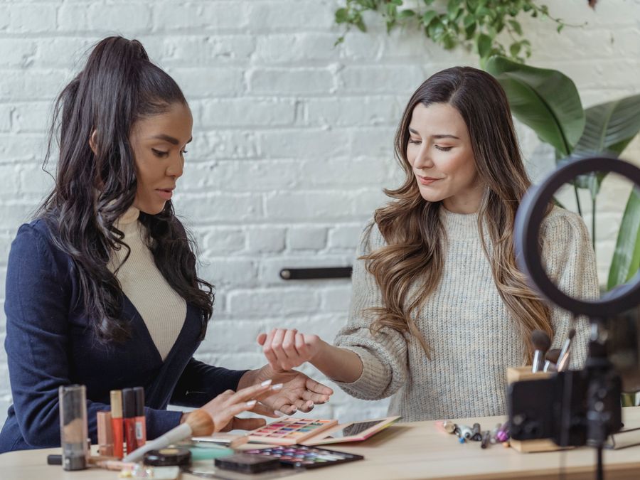 Young positive multiethnic female bloggers testing makeup products while sitting at table and recording vlog on smartphone placed on tripod with ring light
