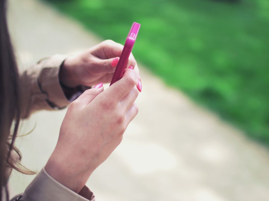 Woman holding a pink smartphone with both hands in a casual outdoor setting.