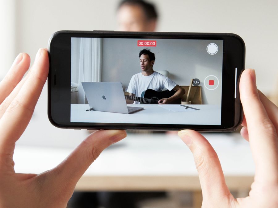 Man playing guitar while recording a vlog video using a smartphone.