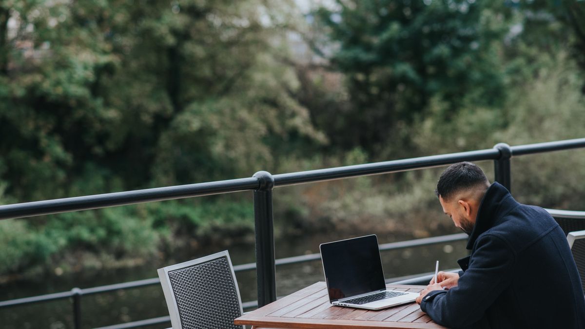 Side view of unrecognizable ethnic male executive taking notes near netbook at cafeteria table on veranda