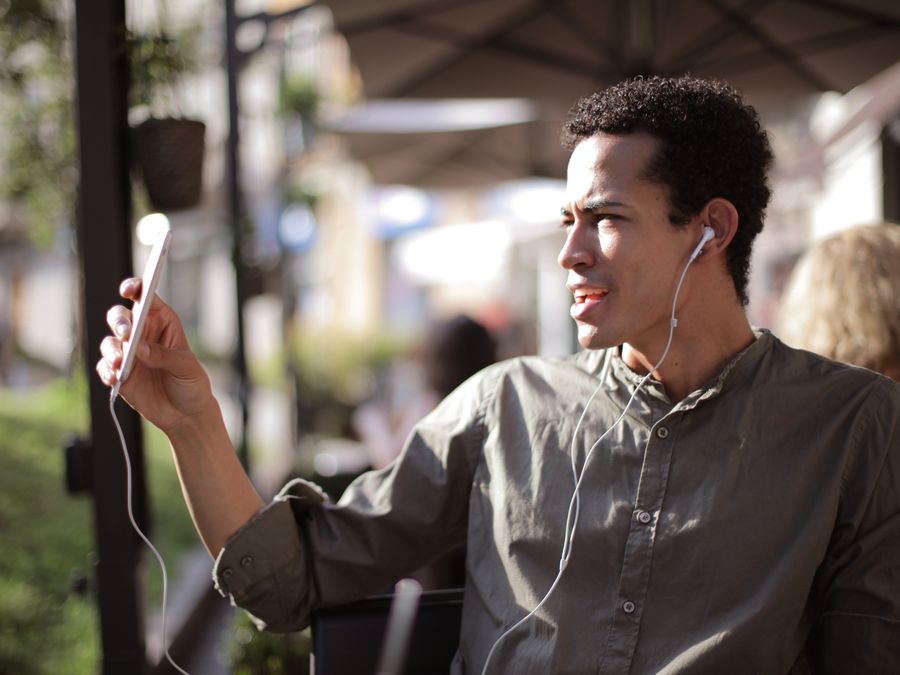 Side view of black male wearing summer shirt using smartphone for video chatting and sitting on outdoor terrace while communicating on social media