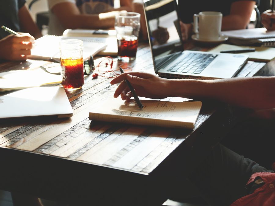 A group of adults engaged in a collaborative office meeting, discussing ideas with notes, laptops, and refreshments.
