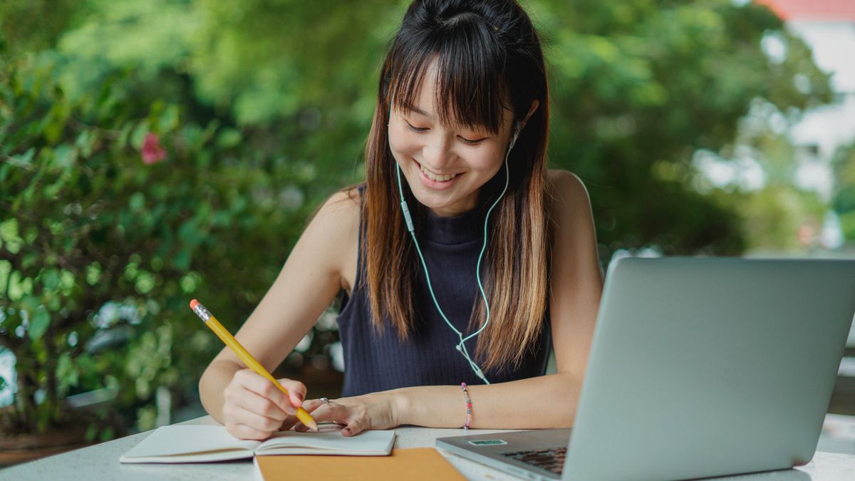 Positive young Asian female student with earphones writing in copybook while doing homework at table with laptop in street cafeteria