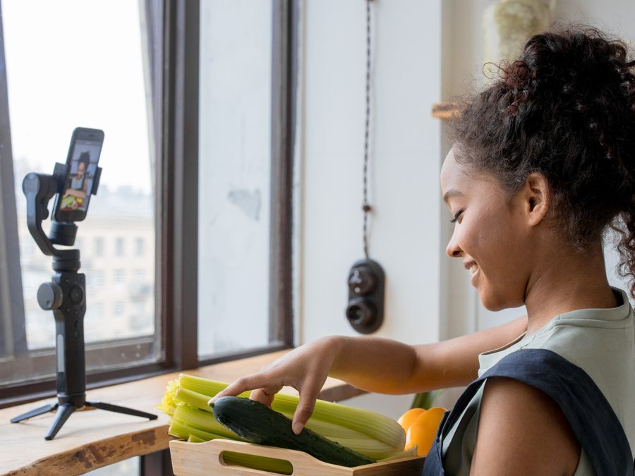 A woman arranges vegetables while recording a cooking video on her smartphone indoors.