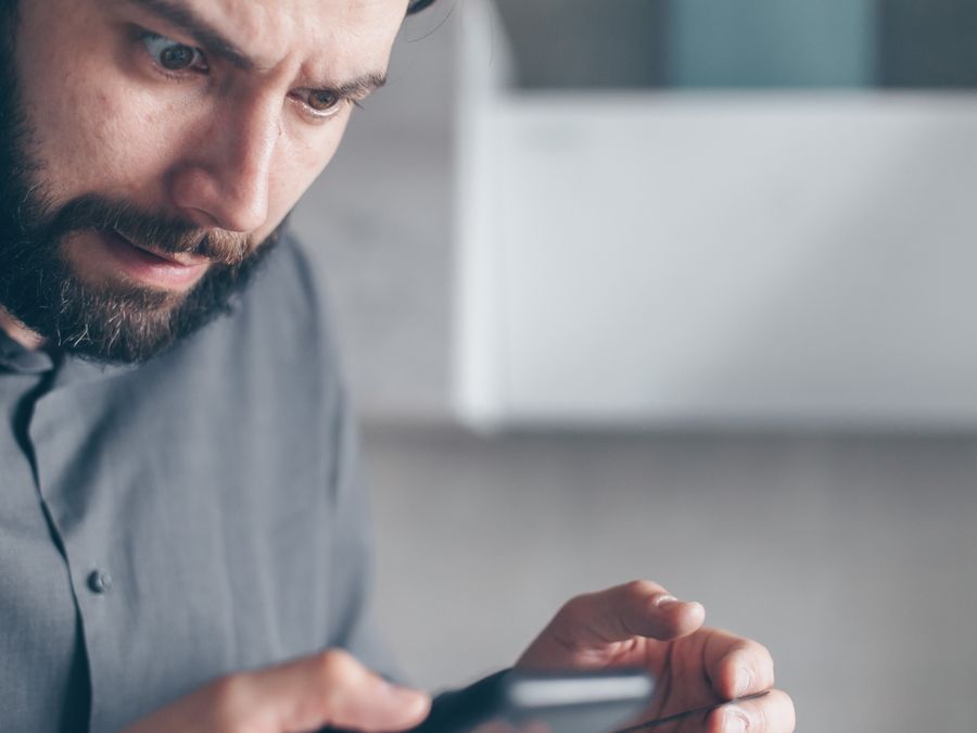 A bearded man in a gray shirt looks shocked while holding a smartphone indoors.
