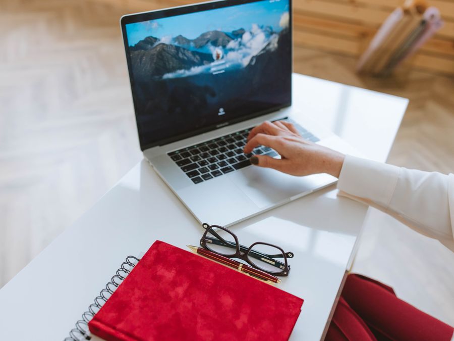 A person working on a laptop with a red notebook and glasses on a white table.
