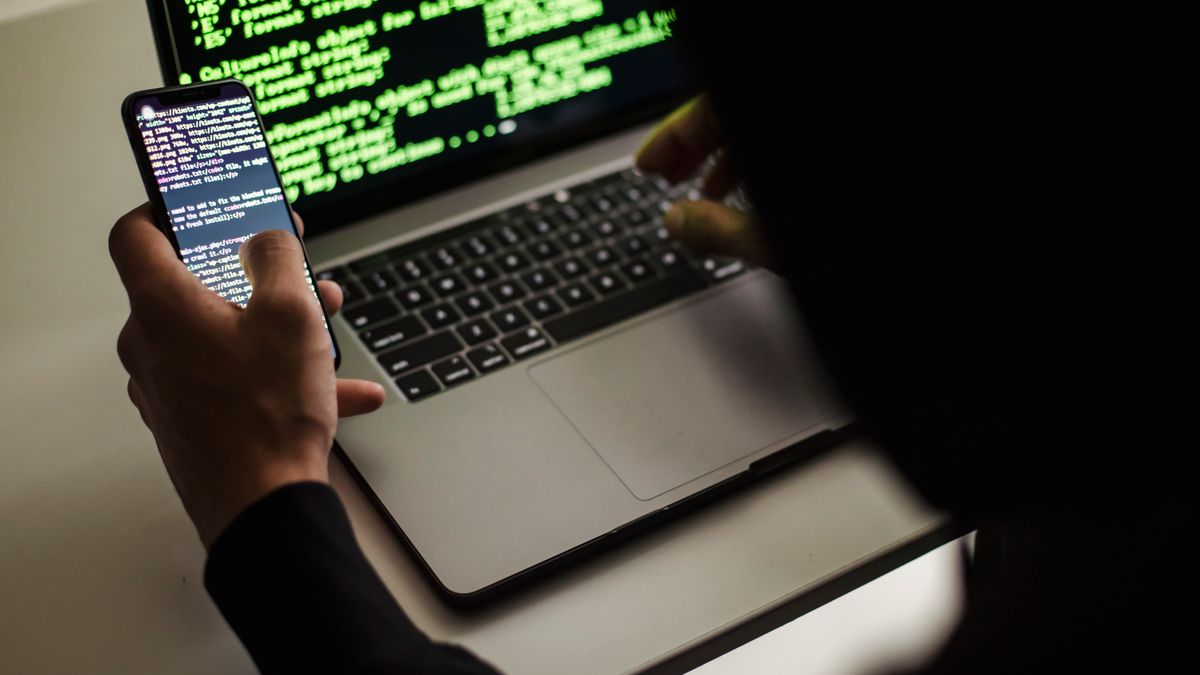 From behind crop male programmer in black hood browsing netbook and mobile phone while working in office