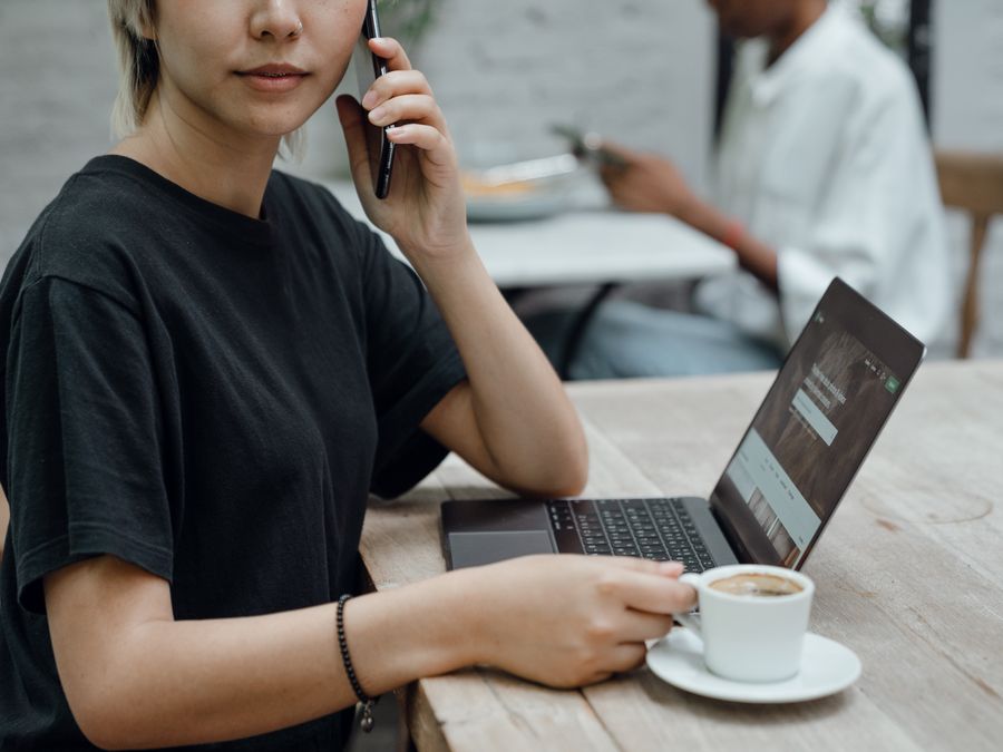 Crop Asian lady in black tee making phone call while sitting at table with coffee and laptop in cafe